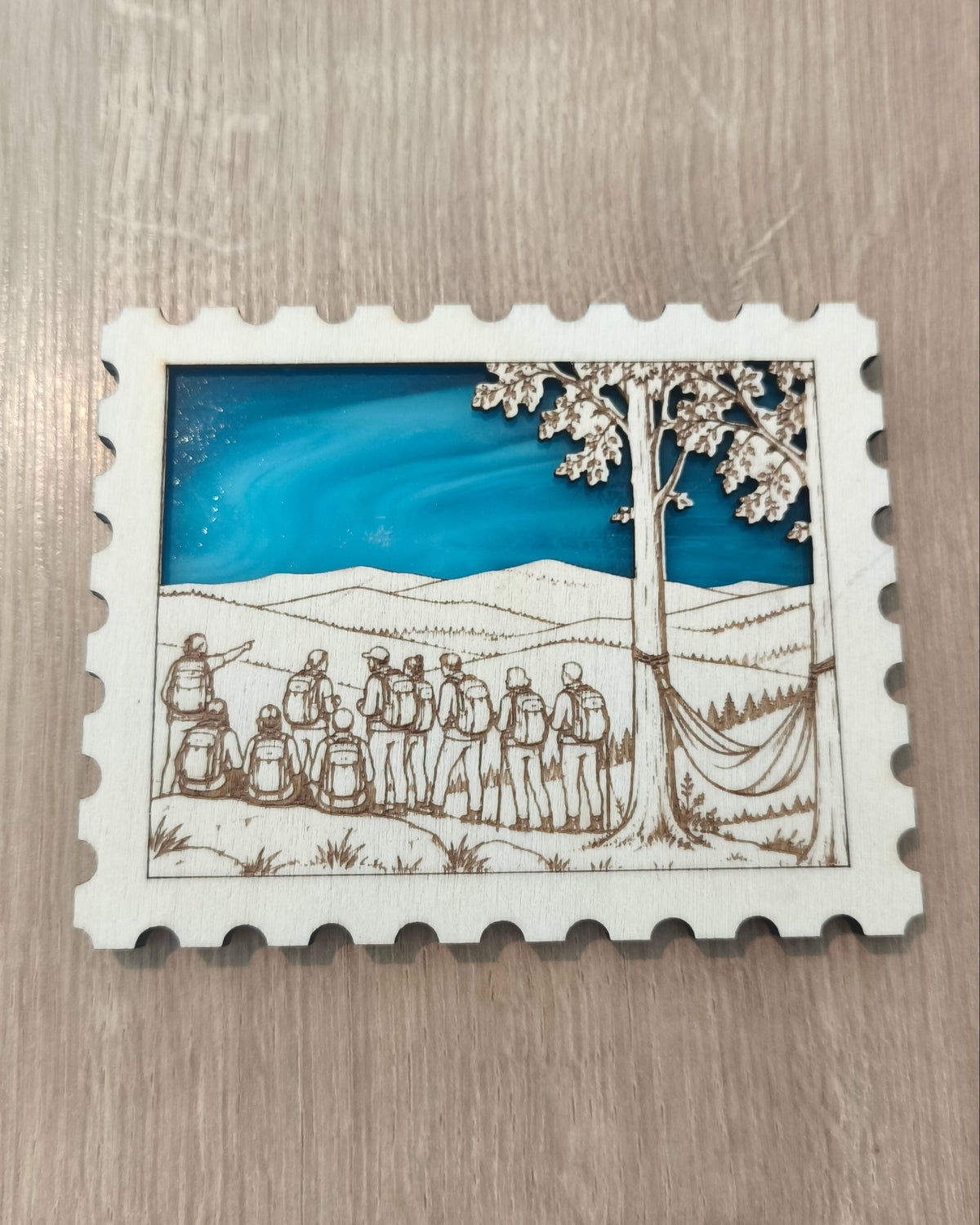 Aimant personnalisé en verre et en bois en forme de timbre représentant un groupe de randonneurs face à un paysage montagnard, et un hamac tendu entre 2 arbres. 