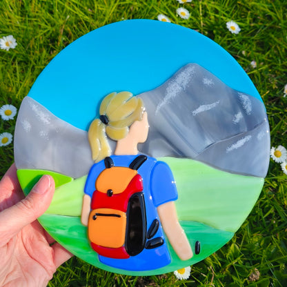 Décoration ronde représentant une randonneuse face à un paysage de montagne en verre fondu.